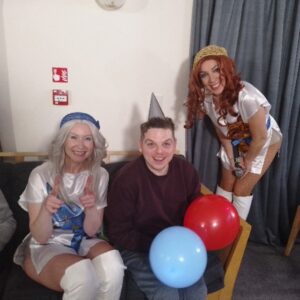 Two people dressed as the two women in ABBA pose with a person in the middle with a party hat on, holding balloons. 