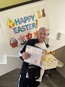 A person holding a drawing and Easter basket filled with treats in front of a colourful sign that reads "Happy Easter."
