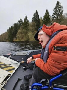Person in red puffer jacket and life jacket is laughing and holding a fishing rod while on a boat. There are trees and a reservoir in the background. 