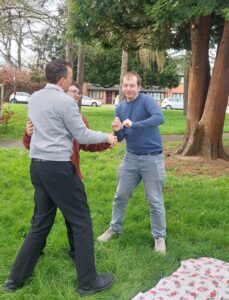 Photo description: Three people dancing in a circle outside on the grass. There is a picnic blanket in the corner of the photograph and trees in the background.