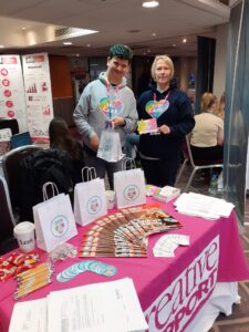Lewis and another person stood behind a Creative Support stall with merchandise laid out on a pink tablecloth.
