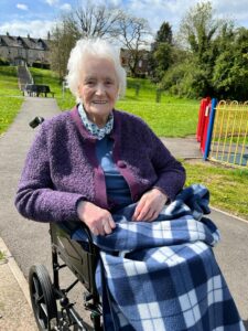 A photo of Sheila, sitting in her wheelchair in a park on a sunny day